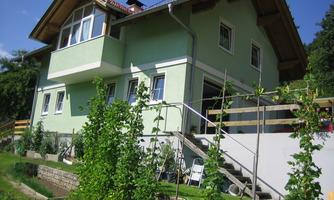 A modern, green house with a balcony and windows. The garden is arranged with plants and a small path.