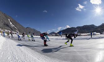A group of speed skaters is racing on a frozen track. In the background, snow-covered mountains and a blue sky are visible.