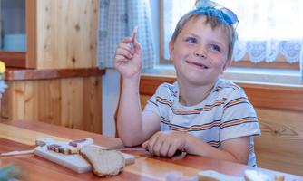 A smiling boy is sitting at a table holding a snack in his hand. In front of him are slices of bread and various spreads.
