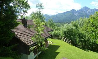 A beautiful wooden house surrounded by green trees and a vast meadow. In the background, majestic mountains and a clear blue sky can be seen.