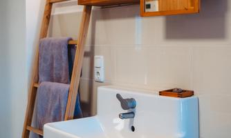 A modern bathroom with a white sink and a wooden shelf. Towels hang on a practical ladder next to the sink.