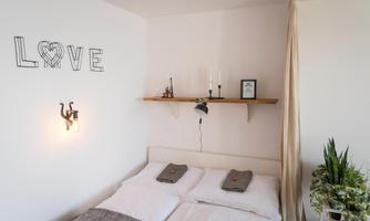 A cozy bedroom with a double bed and decorative pillows. On the wall, it says "LOVE" and there is a small shelf with candles and plants.