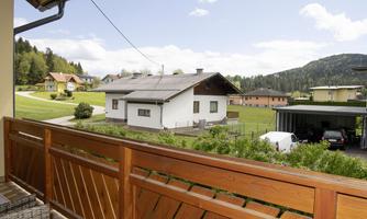 A quiet rural setting with several houses and a green garden. In the background, gentle hills and a blue sky can be seen.