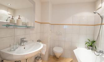 A modern bathroom with white tiles and an elegant bathtub. There is a washbasin with a mirror and plants.