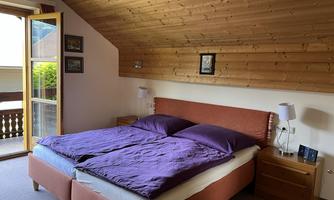 A bright bedroom with a double bed and purple bedding. A balcony door and wooden paneling give the room a cozy atmosphere.