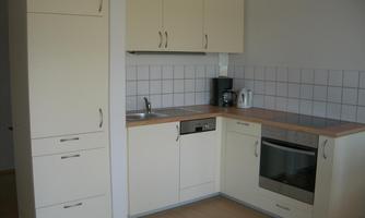 A modern kitchen with light cabinets and a wooden countertop. It is equipped with a stove, a sink, and a coffee machine.