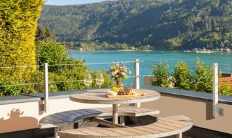 A beautiful terrace with a round table, on which wine and fruit are placed. In the background lies a clear lake and green mountains.