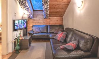 A cozy living area with two leather sofas and a television. The room has a wooden ceiling and large windows with curtains.