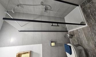 A modern bathroom with a shower and a toilet. The walls are tiled in gray, and there is a small wooden shelf above the shower.