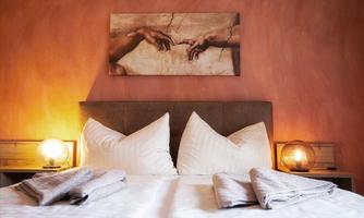 A cozy bedroom with a large bed and two pillows. A painting depicting hands hangs on the wall, and there is soft light from two bedside lamps.