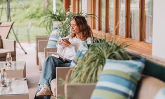 A woman is sitting relaxed on a couch and enjoying a snack. Around her are green plants and bright cushions.