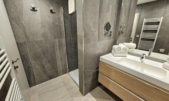 A modern bathroom with a shower, a large sink, and wooden shelves. The walls are designed in dark stone, and towels are neatly arranged.