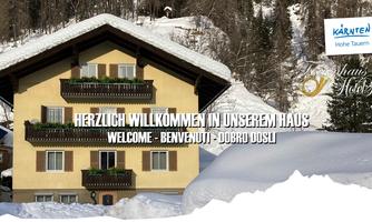 A chalet in the snow with several balconies and an inviting welcome sign. Snow-covered mountains are visible in the background.