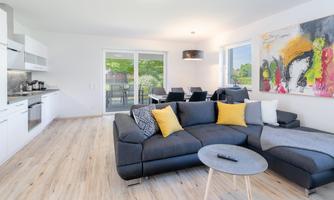 A modern living room with a gray sofa and colorful cushions. The room is bathed in light and has large windows overlooking the outdoors.