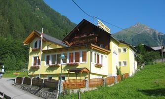 A charming yellow house in the mountains with a balcony and flower boxes. Surrounded by green trees and a clear blue sky.