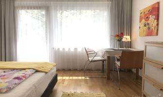 A bright, modern room with a bed and a desk. Large windows provide plenty of natural light.
