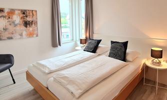 A modern bedroom with a double bed and stylish decoration. The room has large windows and a peaceful atmosphere.