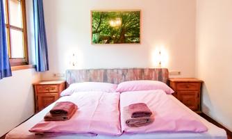 A cozy bedroom with a double bed and fresh bedding. Windows and a picture of a tree are hanging on the walls.