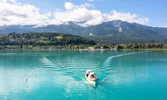 A boat sails over the turquoise blue water of a lake, surrounded by green hills and mountains. The sky is clear and sunny with a few clouds.