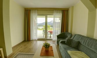 A cozy living space with a green sofa and a table. The large window front offers a view of the mountains and is adorned with airy curtains.