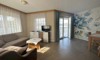 A bright living room with a cozy couch and a television. Large windows offer a view of nature, and a flower wall gives the room a friendly atmosphere.
