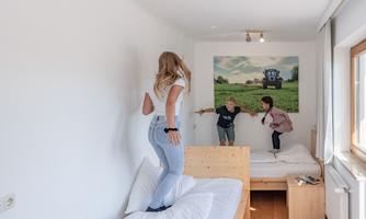 Three children are playing in a bright room. Two children are standing on a bed, while the third one is smiling and standing by a wall.