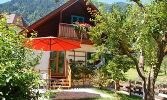 A cozy wooden house with a balcony and a red sun umbrella. Surrounded by a green garden and mountains in the background.
