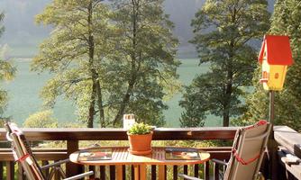 A cozy balcony with two chairs and a table. In the background, there are trees and a calm lake.