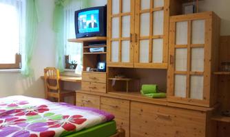 A cozy bedroom with a wooden bed and a colorful duvet cover. In the room, there is a television and a desk, surrounded by bright curtains.