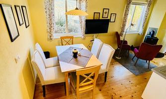 A cozy dining area with a wooden table and white chairs. Bright walls and a large window create an inviting atmosphere.