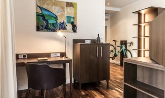 A modern room with a desk, a chair, and a dresser. A colorful piece of art hangs on the wall, and a bicycle is in the room.