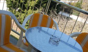 A balcony with a round table and two chairs in yellow-orange stripes. In the background, green trees can be seen.