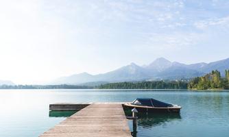 A picturesque lake with a wooden pier and a small boat. In the background, gentle mountains and a clear sky can be seen.