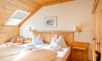 A cozy bedroom with a wooden ceiling and two single beds. Bright walls and windows create an inviting atmosphere.