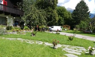 A beautiful garden with colorful flowers and a well-maintained lawn. In the background, there is a residential house and a delivery vehicle, surrounded by trees.