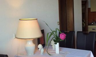 An elegant table with a white tablecloth, a lamp, and a vase of flowers. In the background, chairs and a room are visible.