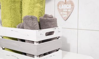 A wooden box with green and gray towels sits on a bathroom shelf. A heart-shaped sign hangs on the wall with the inscription "LAUGH EVERY DAY OF YOUR LIFE".