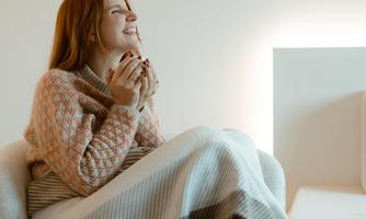 A smiling woman sits in a cozy armchair. She holds a cup in her hands and is wrapped in a blanket.