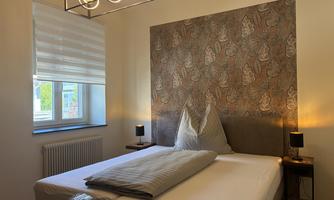A modern room with a bed and an elegant light. The walls feature an artistic pattern, and a window provides daylight.