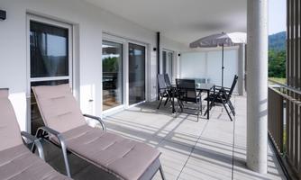 A modern terrace with lounge chairs and a table with chairs. In the background, trees and gentle hills can be seen.