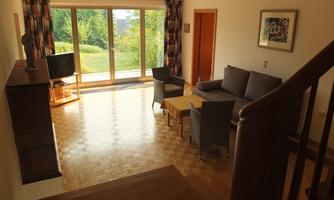 A bright living room with a sofa, a table, and a television corner. Large windows offer a view of the garden.