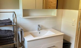 A modern bathroom with a white sink and a mirror cabinet. Towels are neatly stacked on a shelf, and the floor is tiled.