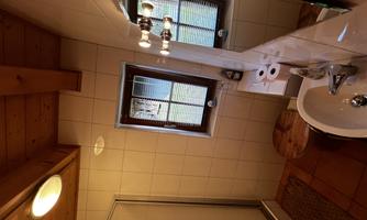 A small, bright bathroom with tiles and wood details. There is a shower, a sink, and a window that lets in daylight.