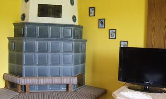 A traditional tiled stove in a light yellow room. In the background, a television and some framed pictures can be seen on the wall.