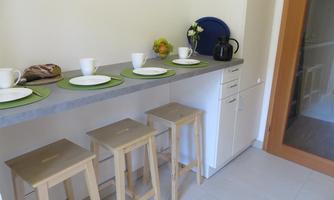 A modern kitchen with a long countertop and four wooden bar stools. On the countertop, dishes, a coffee pot, and fresh fruit are decorated.
