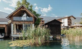 A beautiful house by the water with a porch and green plants. In the background, there are more buildings and a blue sky.