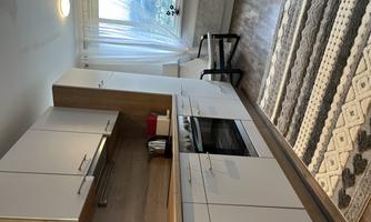 A modern kitchen with white cabinets and wooden details. In the foreground is a patterned rug, and a cozy armchair sits next to the window.