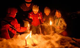 A group of children stands in the snow holding torches in the darkness. They are smiling and enjoying the wintry atmosphere.