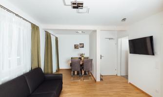 A modern living room with a black sofa and a dining table for four people. The walls are light and there are large windows with curtains.
