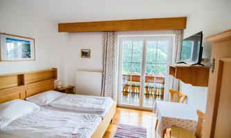 A bright bedroom with a double bed and a table. Large windows offer a view of a balcony.